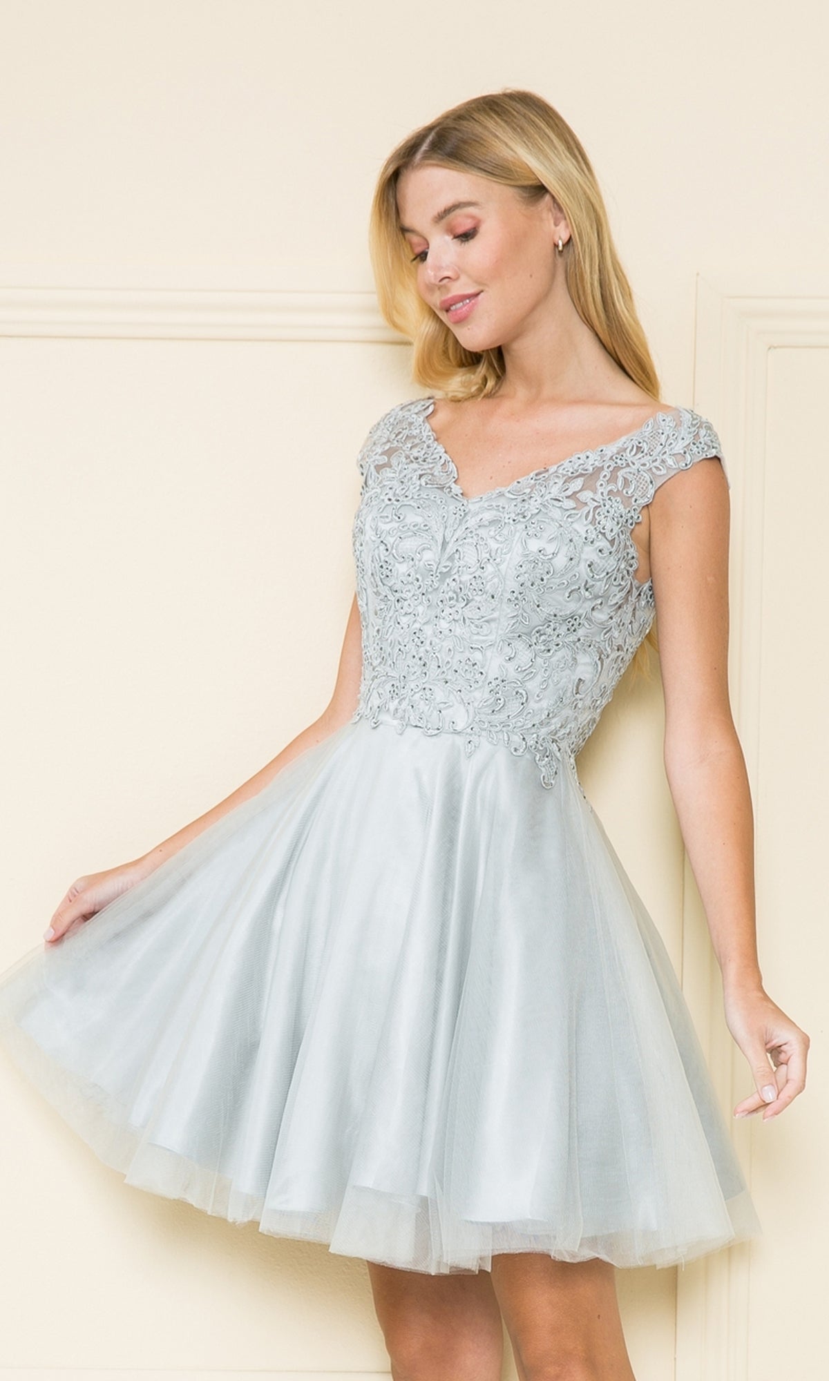 A woman in a silver Lace-Bodice Short Babydoll Homecoming Dress 8902 with lace detailing on the bodice and cap sleeves stands against a beige background.